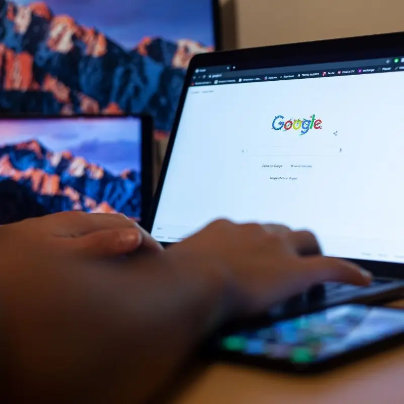 person typing on a laptop with google search page open, with monitors showing mountain scenery in the background